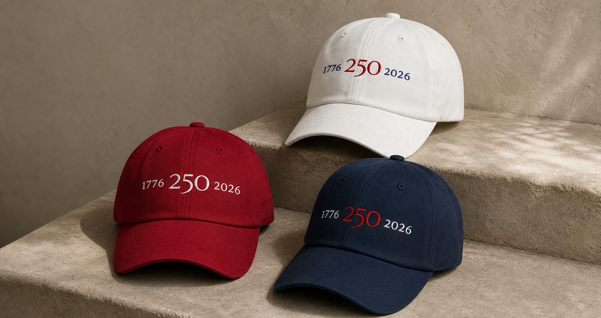 White, red, and navy 1776–2026 embroidered hats from Atlas Stitch arranged on a warm neutral stone background.