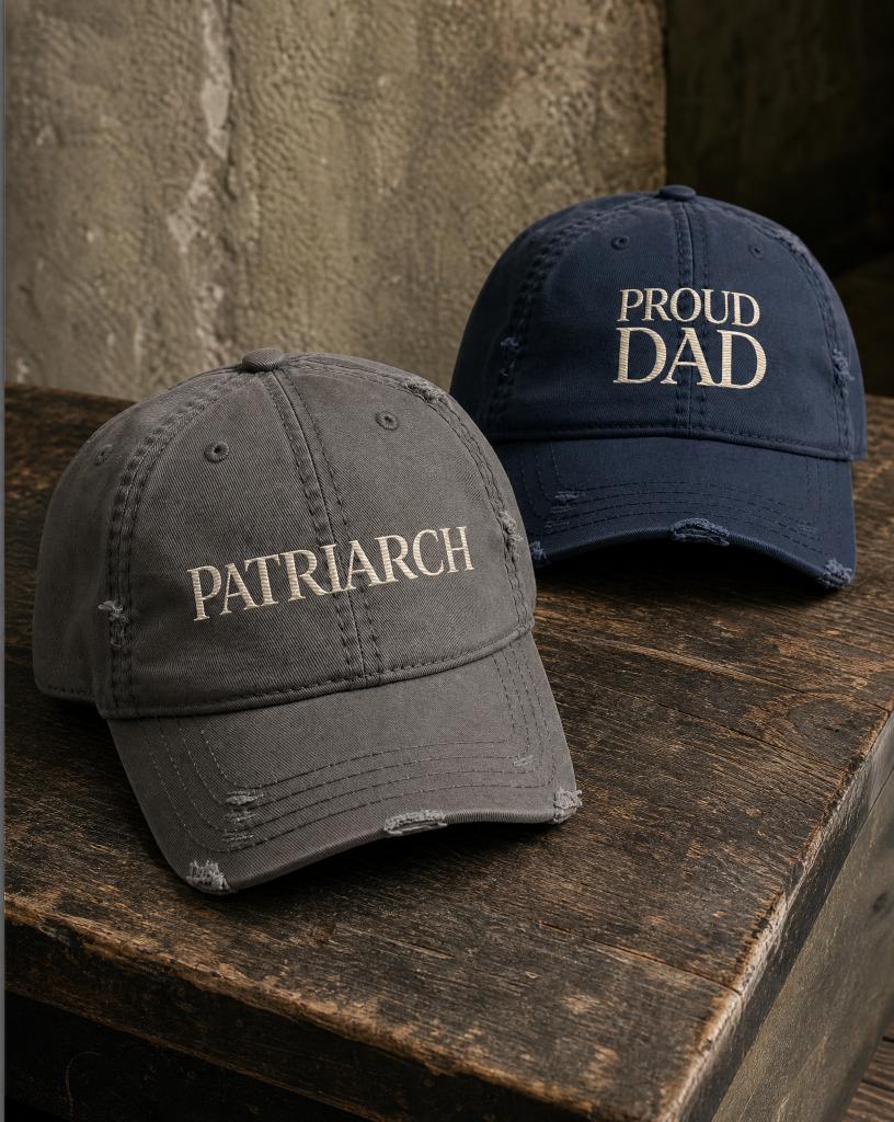 Charcoal PATRIARCH embroidered hat and navy Proud Dad embroidered hat from the Atlas Stitch Father Capsule on a dark wood surface.