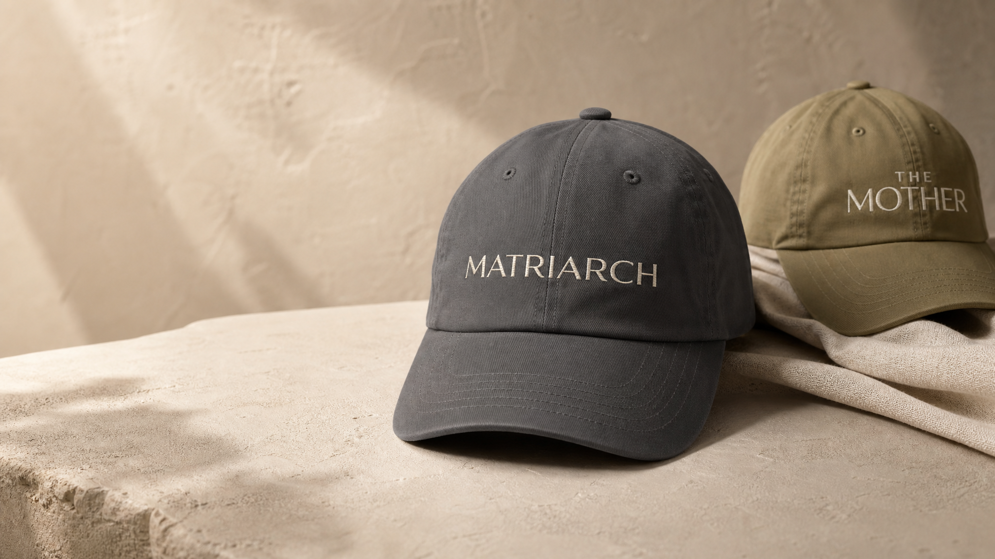 Charcoal MATRIARCH embroidered cap and olive THE MOTHER embroidered cap from the Atlas Stitch Mother Capsule on a warm neutral background.